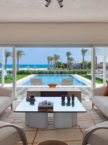 White living room with glass coffee table opens through sliding doors to pool, beach and sea.