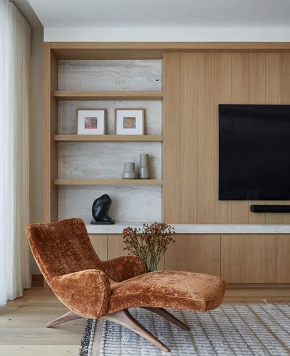 Orange velvet chaise lounge chair in front of oak paneled wall with stone-backed shelving niche and mounted tv