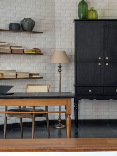Study nook with wooden desk, cane chair, floating book shelves and a black armoire against a brick wall