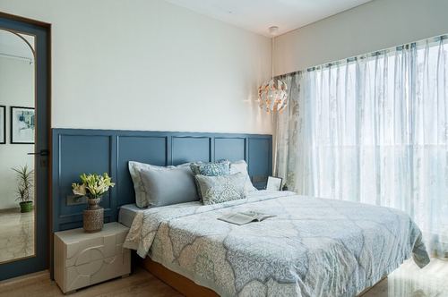 Bedroom with blue panelled headboard wall, patterned bedding, and a geometric pendant light beside sheer curtains