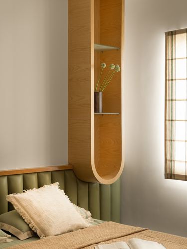 Curved wooden wall niche with vase beside olive channel-tufted headboard in sunlit bedroom