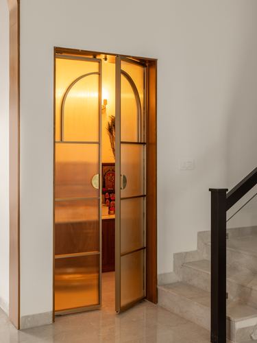 Open arched brass-framed glass door revealing a warmly lit pooja room beside a staircase