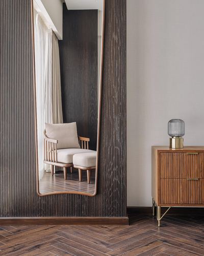 Fluted wood wall with tall mirror reflecting an armchair, beside a wooden chest of drawers and lamp