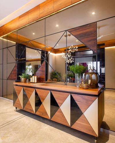 Wooden console with triangular mirror-inlay front beneath a mirrored geometric wall panel and chandelier