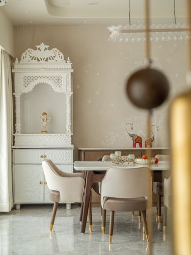 White carved pooja mandir beside a dining table with cushioned chairs and brass accents