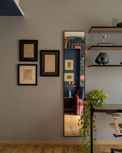 Grey wall with framed sketches beside a floor mirror reflecting a tropical bedroom, shelves and desk to the right
