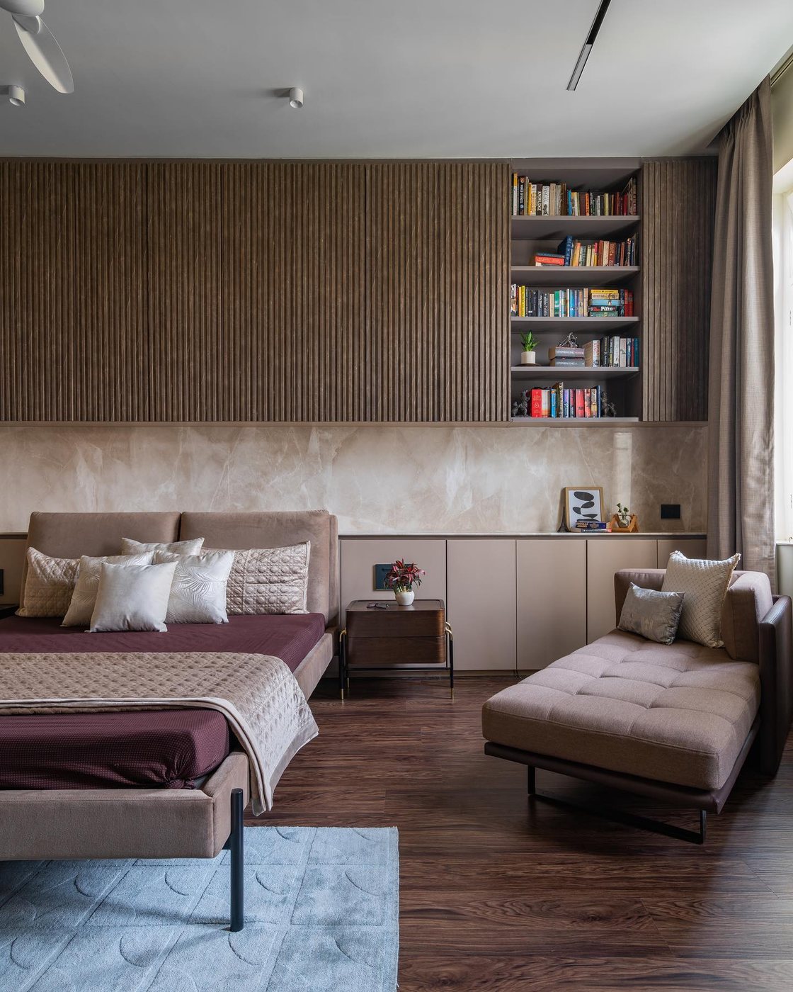 Bedroom with fluted wood wall, built-in bookshelf, maroon bed and tufted chaise lounge