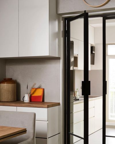 View through black steel-framed glass door into a white kitchen with wooden counter and ceramic jar in foreground