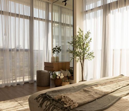 Sunlit bedroom corner with sheer curtains, wooden dresser, mirror, and potted olive tree