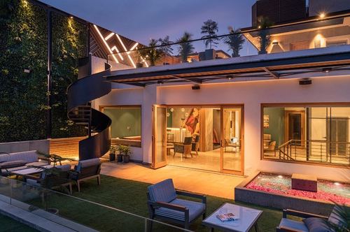 Twilight view of a Delhi rooftop terrace with spiral stair, green wall, lounge seating, and glowing interior beyond glass doors