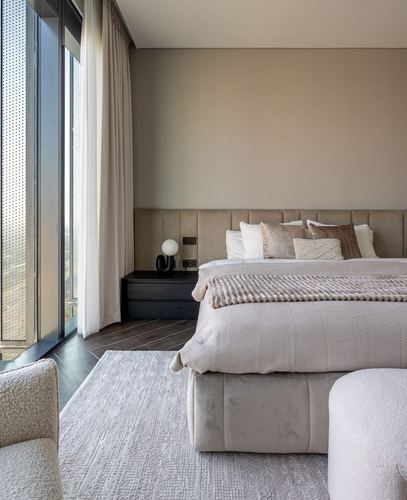 Neutral toned bedroom with channel-tufted headboard, dark nightstand, and floor-to-ceiling sheer curtains beside a city view.