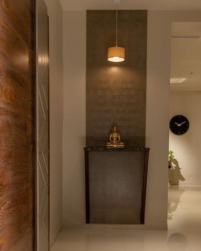 Small pooja nook with engraved mantra panel, pendant light, and brass buddha figurine on metal console