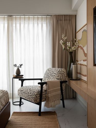 Bedroom corner with patterned armchair by sheer curtains, side table, and floating wood console with art