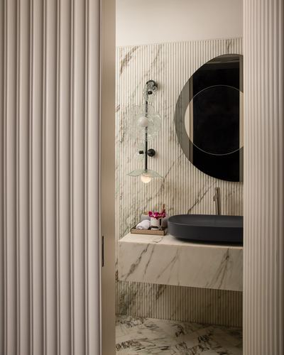 Marble powder room seen through fluted door with round mirror and dark vessel sink