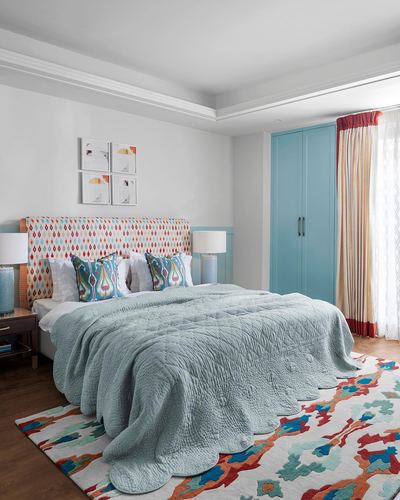 Bedroom with patterned headboard, blue quilted bedspread, colorful rug, and teal wardrobe