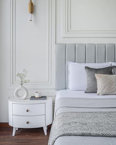 White bedroom corner with tufted grey headboard, round white nightstand, and brass sconce
