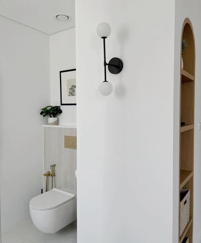 White bathroom corner with black twin-globe sconce, terrazzo floor and arched wood shelving beyond