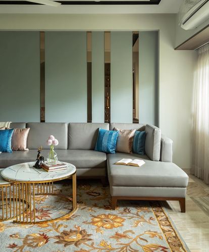 Grey L-shaped sofa with blue cushions against a sage green wall with brass inlays, floral rug below.