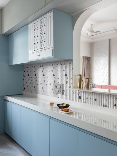 Powder blue kitchen with fluted white backsplash, jali cabinet, and arched opening to living room beyond