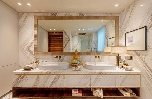 Double vanity bathroom with marble-look tile walls, gold-framed mirror, twin vessel sinks and dark wood open shelving below.