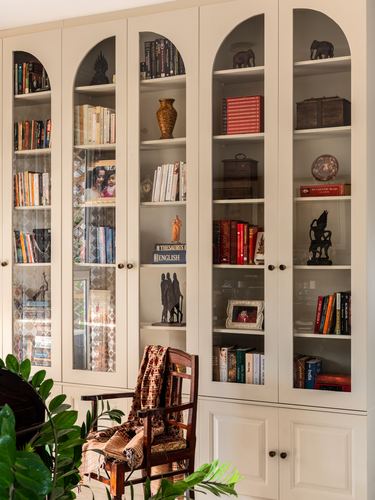 Cream arched glass bookcase filled with books and curios, wooden chair with throw in front, plant leaves at edge