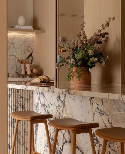 Kitchen island with veined marble countertop, wood cabinetry, bar stools, and a terracotta vase of dried flowers