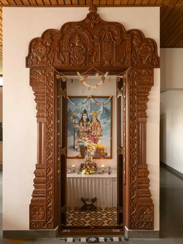 Carved wooden pooja room doorway with deity painting and brass lamps inside