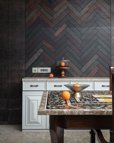 Herringbone wood wall behind white cabinetry with an inlaid stone dining table and oranges