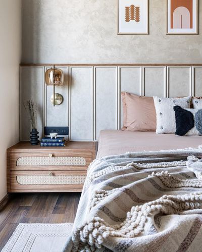 Bedroom corner with cane-drawer nightstand, brass sconce, paneled wall, and layered pink and knit bedding