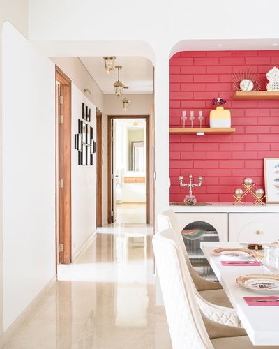 Hallway with framed photos leads to dining nook with pink brick-tile wall and white quilted chairs