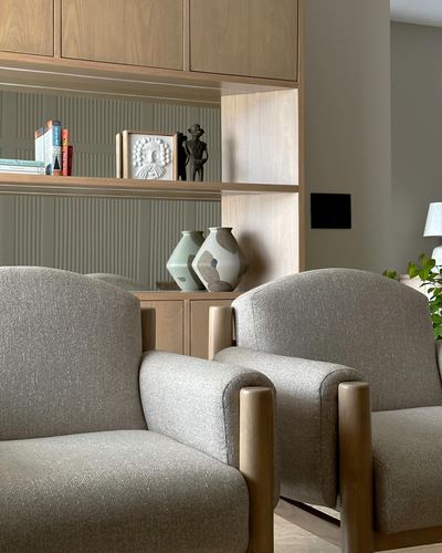Two grey boucle armchairs with wooden legs face a wooden shelving unit displaying ceramics and books