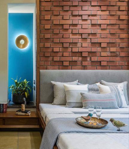 Bedroom with exposed brick headboard wall, grey upholstered bed, and a teal-lit alcove with plant and sconce.