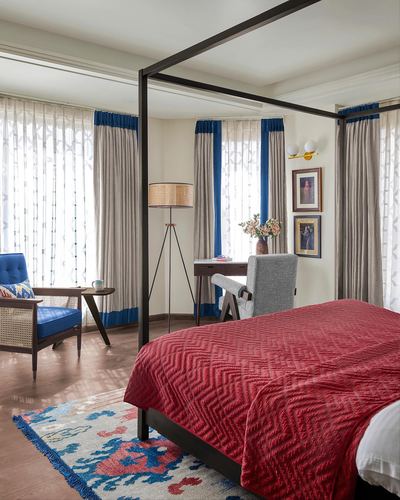 Bedroom with black four-poster bed, red quilted spread, blue-trimmed curtains and cane armchair