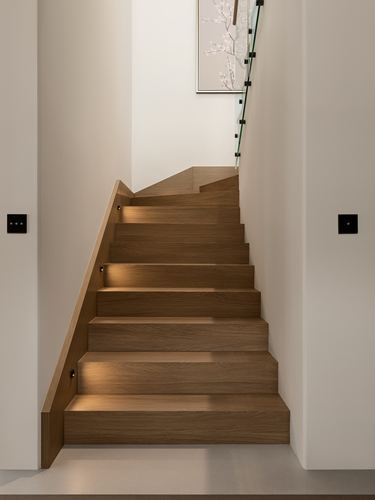 Wooden staircase with glass railing turning upward against a plaster wall with framed art