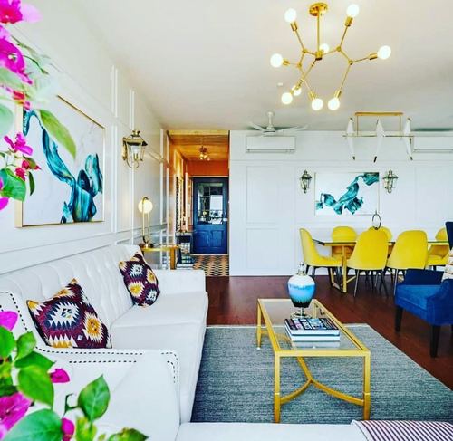 White living room with tufted sofa, gold chandelier, and yellow dining chairs beyond