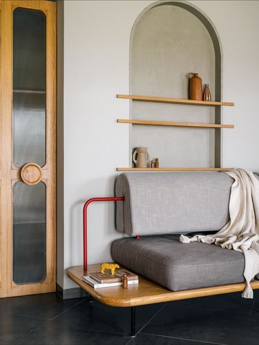 Grey bolster sofa with red metal armrest beneath an arched niche holding wooden shelves and ceramics.