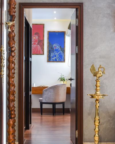View through open door into study with blue and red artworks, velvet chair, brass lamp beside doorway