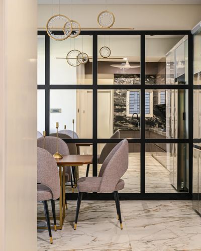 Dining area with mauve chairs and brass pendants seen through black-framed glass partition to kitchen