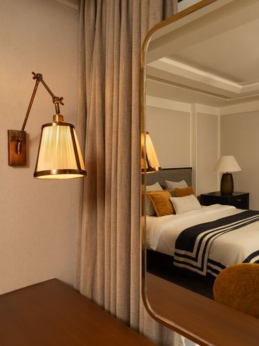 Brass swing-arm sconce beside curtain, mirror reflecting bedroom with mustard pillows and striped throw