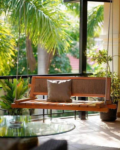 wooden hanging swing with cane backrest on a plant-filled balcony overlooking palm trees