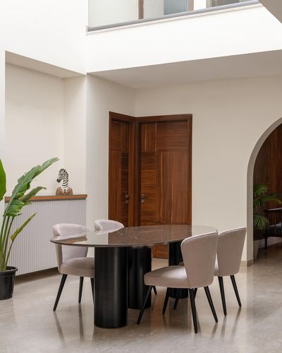Dining area with oval marble-top table, taupe chairs, and wooden panelled corner doors under a double-height ceiling.