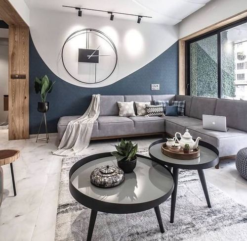 Grey sectional sofa beneath oversized clock on curved blue and white wall, marble floor, nesting round tables