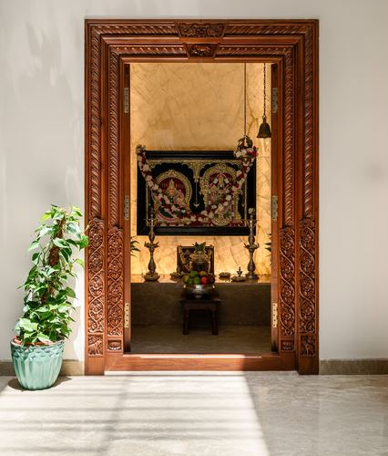 Carved wooden doorway opens to a marble pooja room with hanging bell and deity tapestry