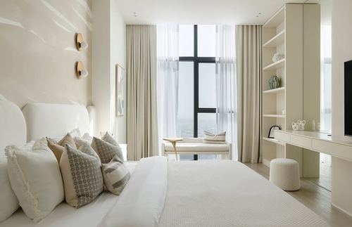 Cream-toned bedroom with upholstered headboard, sheer curtains, floor-to-ceiling window and city view