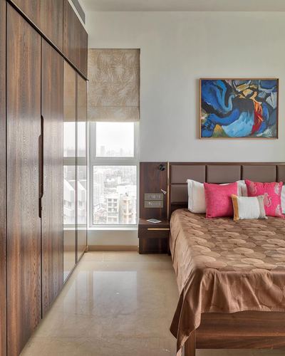 Bedroom with wood wardrobe wall, city-view window, and bed with padded headboard and pink cushions
