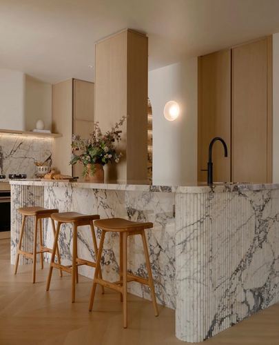 Kitchen island with fluted veined marble waterfall edge, wooden stools, and light oak cabinetry