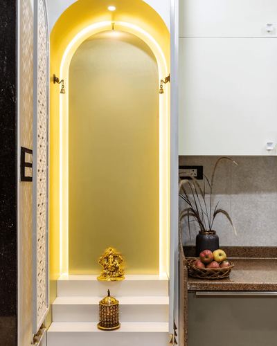 Arched pooja niche lit with warm LED strip, brass bells, deity idol, adjacent kitchen counter