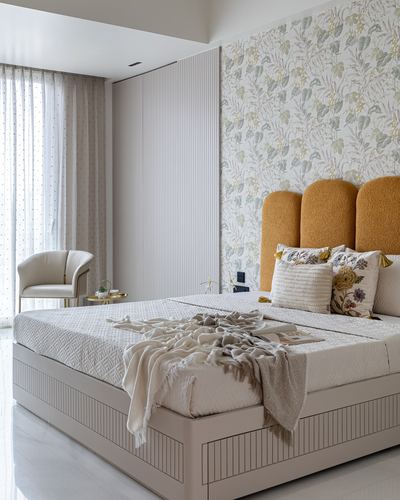 Bedroom with scalloped mustard headboard, floral wallpaper, fluted wardrobe, and cream accent chair by curtained window