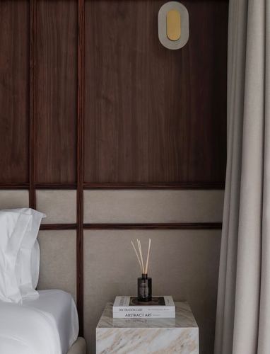 Bedroom corner with dark wood panelling, marble side table holding books and a reed diffuser, beige curtain.