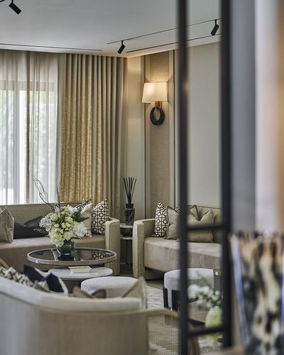 Neutral-toned living room seating area with glass coffee table, floral centerpiece, and sconce lighting, seen through a metal-framed divider.